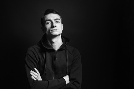 Black and white studio portrait of a young man in a black sweatshirt, arms crossed, seriously looking at the camera, against plain studio backgroundの写真素材
