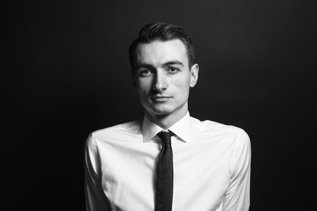 Close up black and white portrait of a young man in a white shirt and black tie, seriously looking at the camera, against plain studio backgroundの写真素材