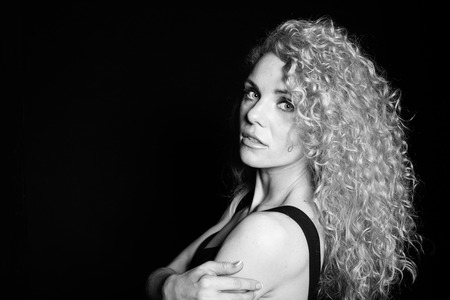 Close up studio portrait of a pretty curly blonde woman, wearing black dress, looking at the camera over the shoulder, against plain studio backgroundの写真素材