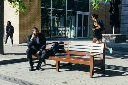 London, UK - July 23, 2018: African ethnicity man speaking on the phone sitting on a bench on a city...