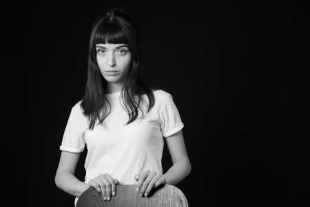 Studio portrait of a pretty brunette woman in a white blank t-shirt, standing behind the chair,...