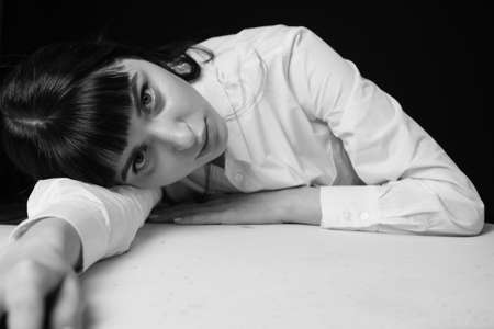 Studio portrait of a pretty brunette woman in a white shirt, leaning on a white table, against a plain black background, looking at the cameraの写真素材