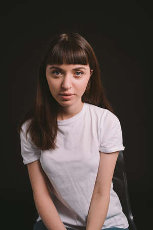 Studio portrait of a pretty brunette woman in a white blank t-shirt, against a plain black...