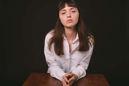Studio portrait of a pretty brunette woman in a white shirt, sitting at the table, against a plain...