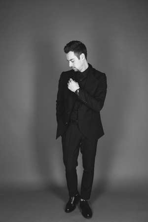 Studio portrait of a young caucasian man in a black blazer, looking down, hand on chest, standing against plain studio backgroundの写真素材