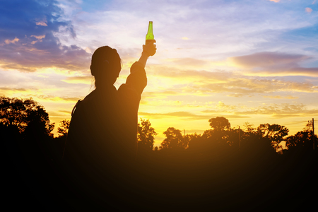 Silhouette woman raised hands holding a green beer bottle on the sunset sky,の写真素材
