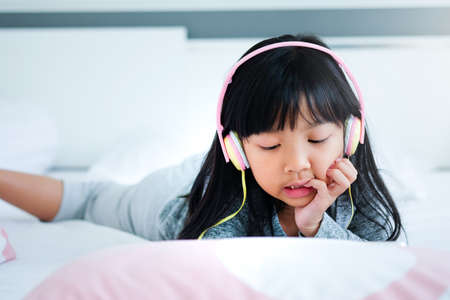 A cute Asian child girl is using headphones to listen to music or Learn in the digital age on a white mattress in her bedroom.の写真素材