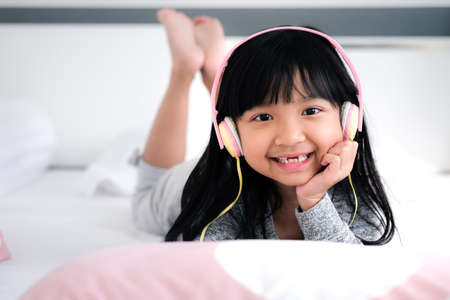 A cute Asian child girl is using headphones to listen to music or Learn in the digital age on a white mattress in her bedroom.の写真素材