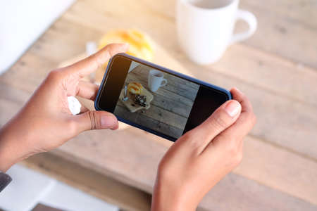 Women's hands are using smartphones, taking bread and coffee before eating for social media.