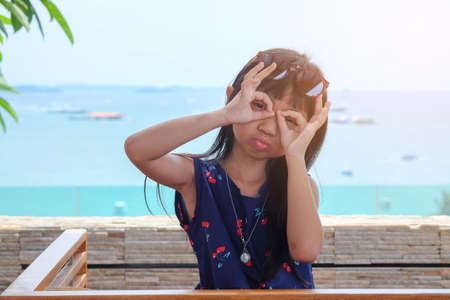Cute asian child girl wearing blue dresses with holding fingers near eyes like glassesの写真素材