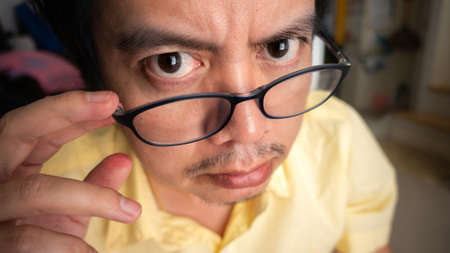 Asian man wearing a yellow shirt is looking under his glasses with suspicion or dazedの写真素材