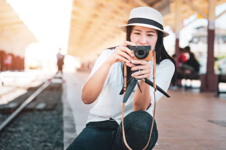 Asian woman tourist wearing a white T-shirt, wearing a hat, holding a camera and taking pictures at the train stationの写真素材