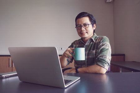 Asian man sitting with a laptop drinking coffee at his deskの写真素材