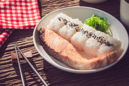 Grilled salmon with soy sauce with Japanese rice topped with black sesame seeds in a black plate on a wooden tableの写真素材