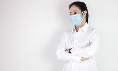 Asian woman in a white shirt standing with arms folded wearing a mask to protect against the corona virus or covid 19 on a white background with copy spaceの写真素材
