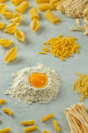 Different types of pasta forming a circle around flour with an egg yolk on a gray background, ready to be cookedの写真素材