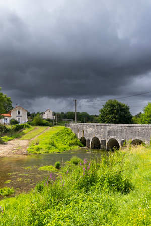 Scenic view of an old stone bridge over a shallow river with houses and lush vegetation under a dramatic stormy sky in Thorey sur Ouche, Bourgogne Franche Comteの写真素材