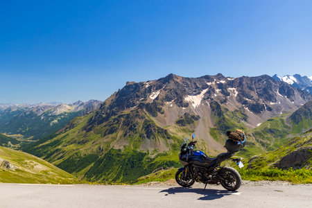 Blue motorcycle parked on asphalt road facing a breathtaking view of the French Alps on a sunny summer dayの写真素材