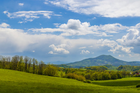 Spring landscape with green fields, trees and Hazmburk Mountain near Ustek, Czech Republic, under partly cloudy skyの写真素材