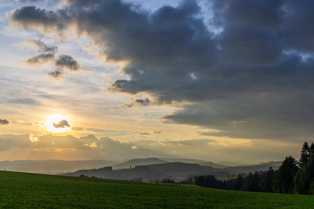 Sun setting behind clouds over rolling hills in Chvalec, Czechiaの写真素材
