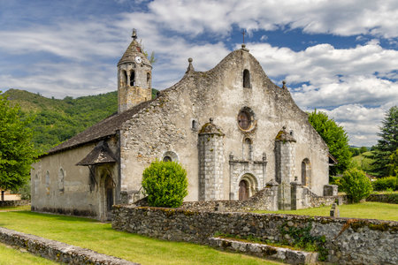 The Church of Saint Jacques in Moulis, Ariege, France, presents its weathered stone facade and bell tower against a backdrop of lush greenery and a partly cloudy skyの写真素材