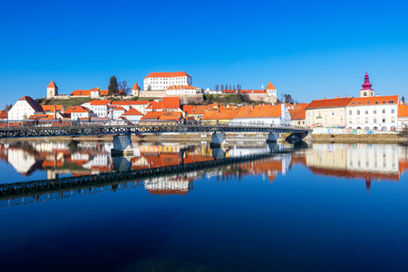 Ptuj Castle and the city reflecting on the Drava River with a pedestrian bridge in the foreground on a sunny dayの写真素材