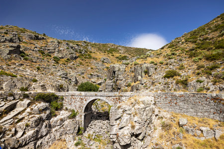 Old stone bridge crossing a gorge in the mountains of Villarejo del Valle, Avila, Spain, under a blue skyの写真素材