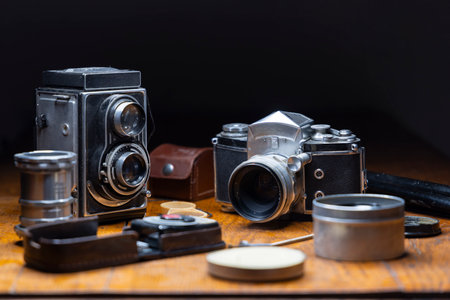 Two vintage cameras and photo equipment resting on wooden table against dark background, representing photography, nostalgia, and collectingの写真素材