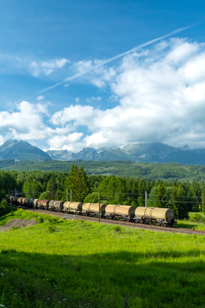 Freight train with tank cars passing through green valley in Slovakia with High Tatras mountains in background on sunny summer dayの写真素材