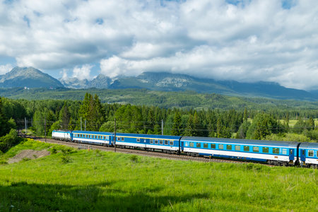 Blue train passing through green fields with High Tatras mountains in background, near Strba, Slovakiaの写真素材