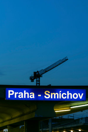 Illuminated Praha Smichov railway station sign contrasting with a towering construction crane silhouette at duskの写真素材