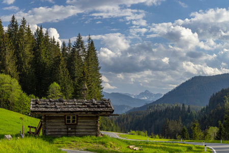 Rustic log cabin hut standing by a winding road in a serene mountainous landscapeの写真素材