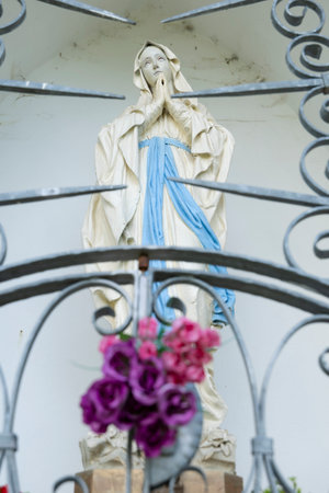 Virgin Mary statue praying behind a metal gate with pink and purple flowersの写真素材