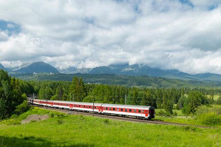 Red and white train moving through green landscape with mountains and clouds in the background