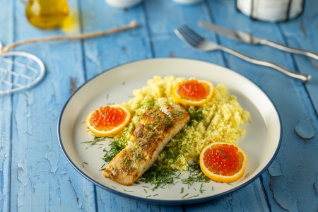 Grilled fish fillet with couscous, dill, lemon slices, and red caviar on a blue wooden table