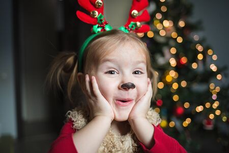 cute funny baby is very surprised. pretty girl in a fancy dress deer on the background of the Christmas tree at home. shocked and humorous kid.の写真素材