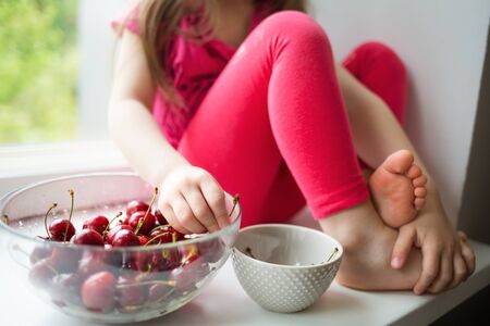 child girl sitting on windowsill at home and eating sweet cherries. summer fruits, healthy seasonal vitamin food.の写真素材