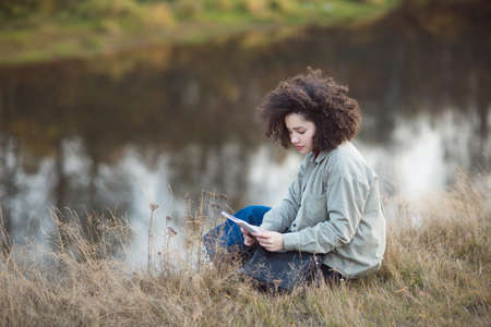 young pretty mixed race teen student girl outdoor in sunlight. happy woman sits on Bank of river reads records in autumn.の写真素材