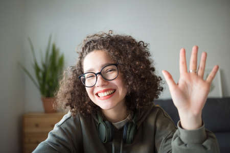 girl looks at camera, waves her hand and smiles. student talks over Internet. Video Conference or...