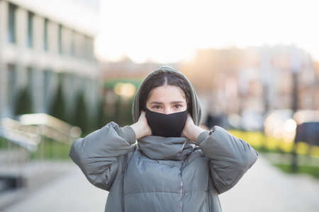 beautiful girl puts on protective mask on city street. woman uses coronavirus protection device in autumn-winter period.の写真素材