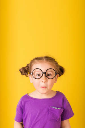 Little cute funny surprised kid child baby girl with glasses in purple T-shirt on yellow wall background. children studio portraitの写真素材