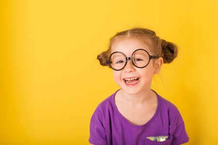 Little cute caucasian funny happy smile kid child baby girl with glasses glasses in purple T-shirt on yellow wall background. children studio portraitの写真素材