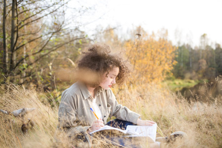young pretty mixed race teen student girl outdoor in sunlight. happy woman sits on Bank of river and writes in notebook in autumn.の写真素材