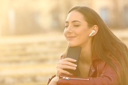Relaxed woman wearing earbuds listening to music on smart phone with closed eyes and a warm light