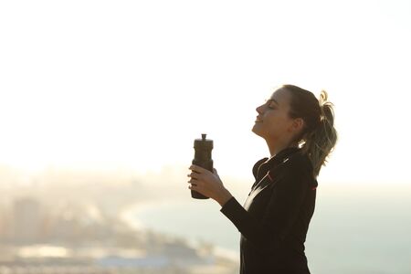 Profile of a runner woman relaxing breathing fresh air holding a water bottle with the city in the backgroundの写真素材