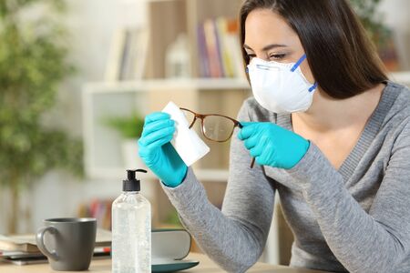Woman with protective mask and gloves disinfecting eyeglasses due coronavirus sitting at homeの写真素材