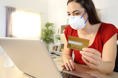 Woman paying online with card on laptop wearing mask in covid-19 confinement sitting on a desk at homeの写真素材