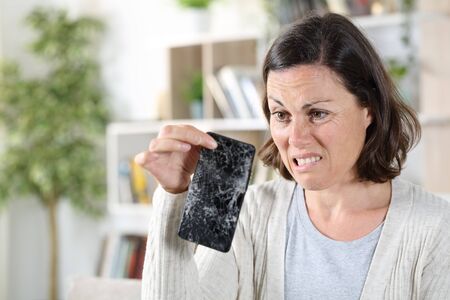 Confused adult woman looking at broken smart phone with shattered screen at homeの写真素材