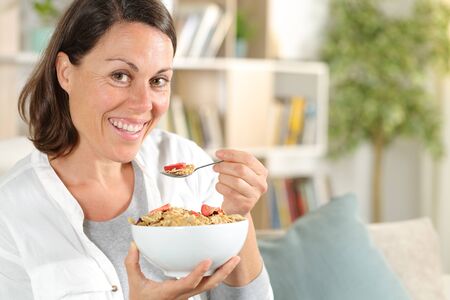 Happy adult woman holding cereal bowl with fruit having breakfast sitting in the sofa at home
