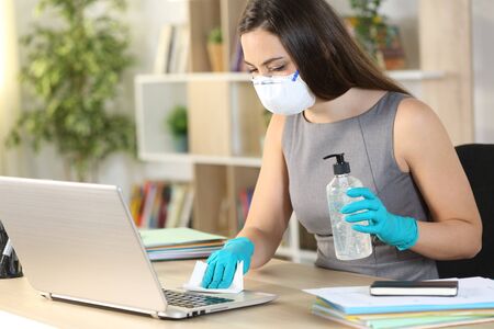 Entrepreneur woman with mask disinfecting computer keyboard with sanitizer and wipe sitting on a...